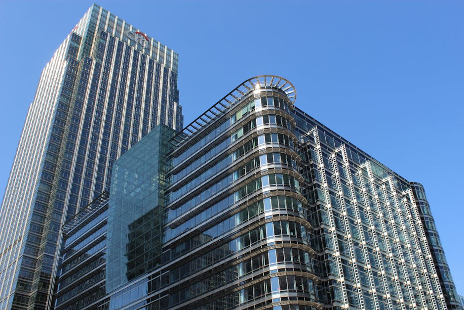 A low-angle view of modern high-rise office buildings with glass facades under a clear blue sky, showing reflections of the surroundings in the windows. The building on the right features curved balco