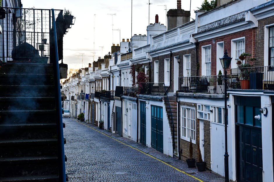 A row of terraced houses with brick facades and white decorative trim on a cobblestone street during daytime. Each property features a small front garden or pathway, with some having plants or shrubs.